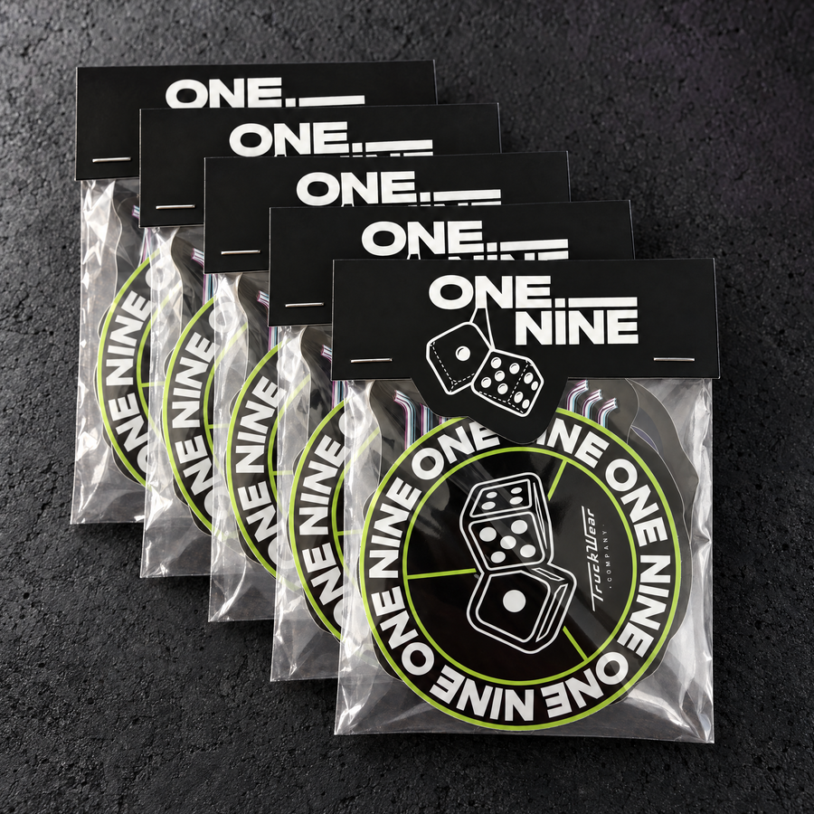 A stack of One Nine Budget sticker packs with short custom top header cards on a charcoal studio surface.