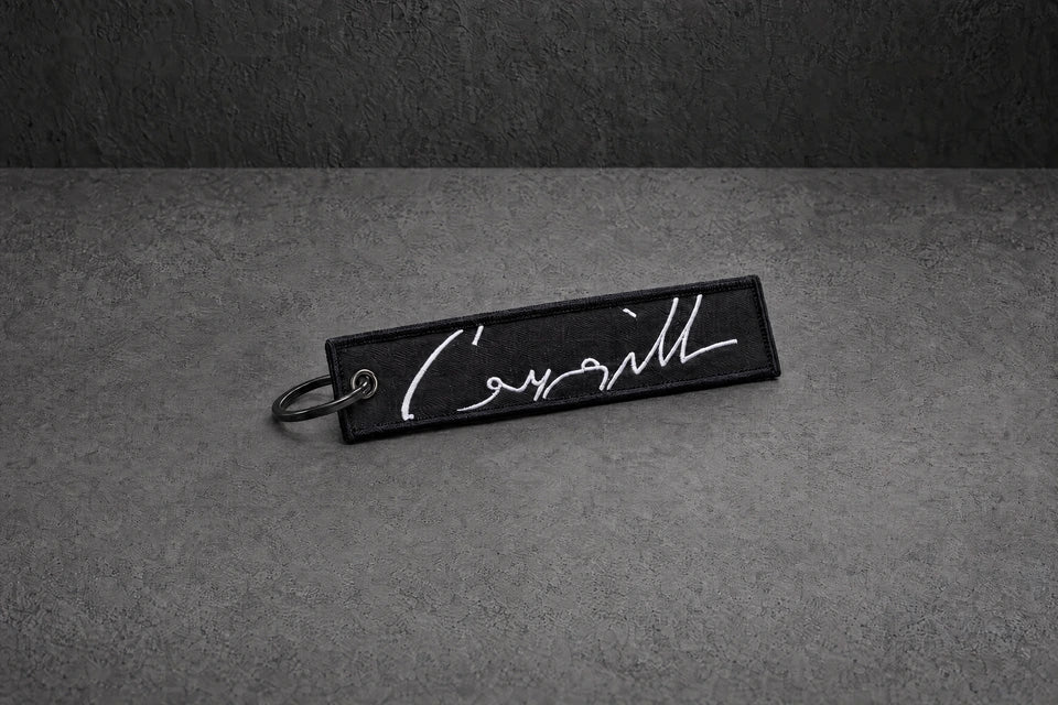 white signature stitched keyring on a charcoal studio surface — Projectprint